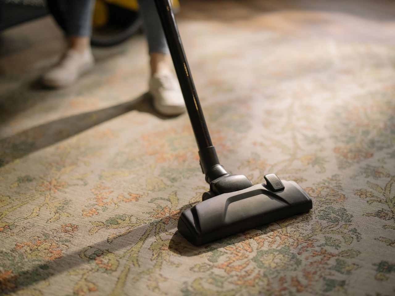 about-02 Close-up of a vacuum cleaner on a patterned carpet in a sunlit room, capturing a moment of household cleaning.